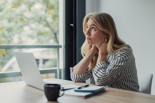 Young adult woman appearing tired and unmotivated, sitting at a desk with a laptop, finding it difficult to focus on work and daydreaming while looking out the window