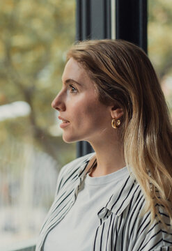 Young woman with blonde hair in a striped shirt gazes out a window in soft daylight, thoughtful and peaceful, capturing a quiet moment of reflection and hopeful daydreaming