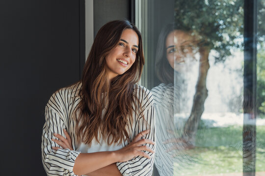 Young contented woman smiling with arms crossed leaning against a window, reflecting lush green garden, representing concepts of contemplation, future, home, well-being, and nature connection