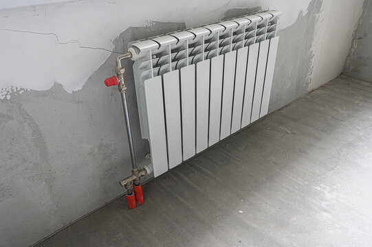 white bimetallic radiator is mounted on an unfinished grey wall with visible cracks; it has a vertical metal pipe and two red shut-off valves, set against a concrete floor