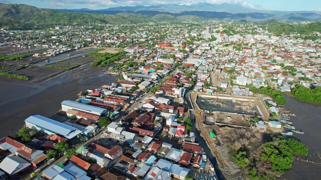 Aerial drone footage of Bima city harbor area at sunset with clouds, in Sumbawa island, Indonesia, with sheds and depots in the front, and the whole city behind, with a main road