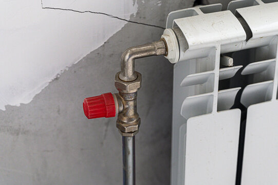 white bimetallic radiator connected to a vertical metal pipe via a curved elbow; a red manual shut-off valve is installed at the junction against a cracked plastered wall