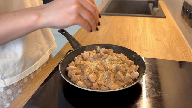 Hand sprinkling black pepper and spices over diced meat in a sizzling frying pan on an induction cooktop. Modern kitchen scene highlighting seasoning process, culinary technique, and flavorful meal 