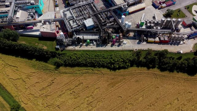 Aerial View of Industrial Complex: An expansive industrial complex with numerous pipes, tanks, and buildings sits adjacent to a cultivated field under a clear sky.