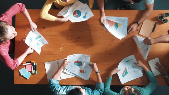 Top view of smart people throw paper and giving high five at meeting table with financial document and stock market analysis paper scattering around. Aerial view of team celebrate project. Symposium.