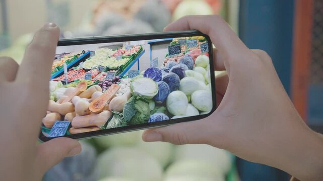 A person takes a photograph of vibrant vegetables in a market, showcasing the variety and freshness available. The scene highlights the appeal of fresh produce and the convenience of modern technology