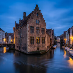 Obraz premium Historic brick townhouse with a stepped gable stands beside a calm canal in Bruges at twilight. Glowing windows reflect on the blue hour water near docked boats and an old stone bridge.