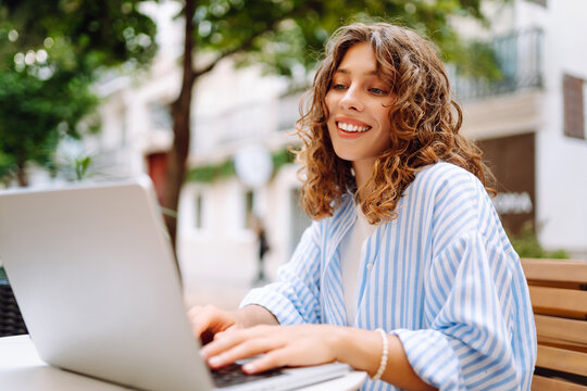 A woman sits at a small table in a street cafe. A happy woman works on her laptop and drinks aromatic coffee on a sunny day outdoors. Concept of freelancing, drinks, and relaxation.