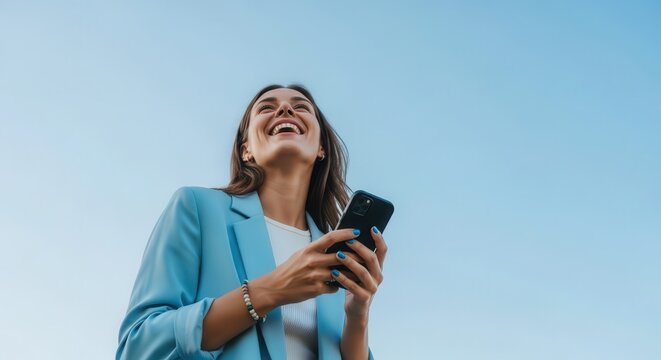 Happy Young Businesswoman Laughing While Using Her Smartphone Outdoors Against a Clear Blue Sky Background