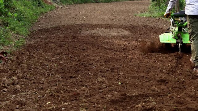 Closeup video of a young Indian farmer plowing agricultural land using a motorized power tiller in rural countryside of Himachal Pradesh, India during summer season.