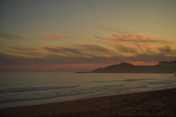 Obraz premium Golden sunset over a misty sandy beach in Portugal’s Algarve region, with soft pink clouds in the sky, a hazy ocean horizon, and distant coastal hills dotted with houses glowing in warm evening light.