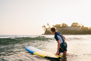 Fototapeta premium focused blonde teenage boy in rash guard holding surfboard while standing in ocean waves on Dewala Beach in Sri Lanka during sunset representing youth active lifestyle tropical travel adventure and wa