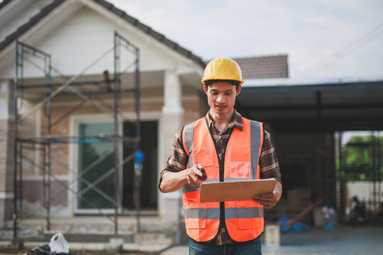 Young Asian construction worker or engineer checking building progress at a residential construction site, wearing hard hat and safety vest