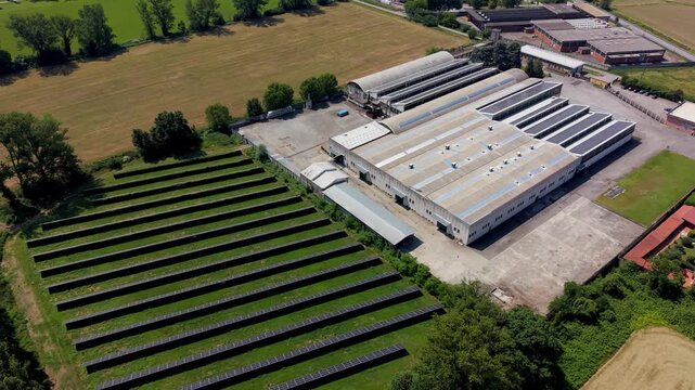 Industrial Park with Solar Panels: An aerial view showcases an industrial park with multiple warehouses and rows of solar panels reflecting sunlight, surrounded by fields and greenery.