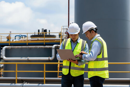 Two Asian male engineers in hard hats and vests look at a laptop. They stand near large industrial tanks and pipes. Planning and inspection occur. They collaborate on a project.