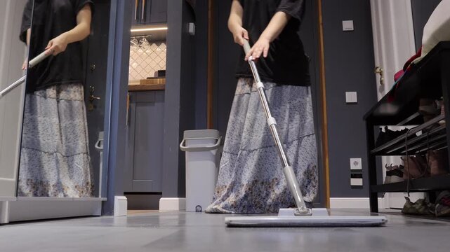 A teenager uses a flat mop near a mirrored closet and gray bucket while doing an entryway household chore.