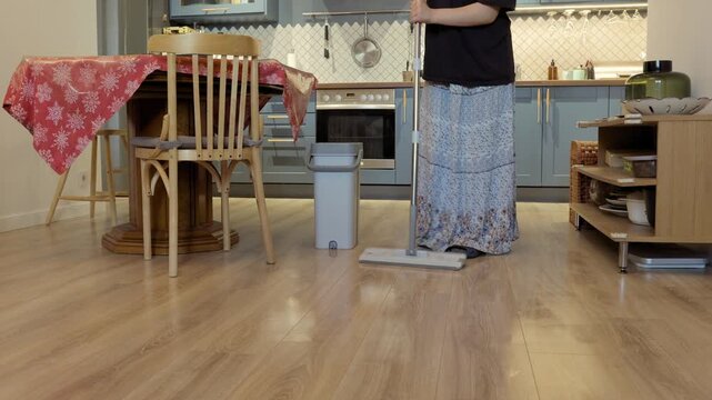A teenager mops a light wooden floor near a dining table and gray bucket during indoor home cleaning.