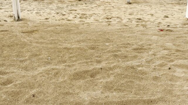 Textured golden sand stretches across a quiet, serene beach. The soft sunlight creates a peaceful atmosphere over the uneven terrain. Tiny shells and footprints dot the warm coastal landscape.