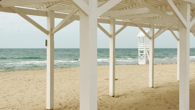 A white wooden structure stands tall on a peaceful, sandy shore. Gentle waves wash against the coast under a soft sky, creating a calm mood. A lonely lifeguard tower waits in the distance.