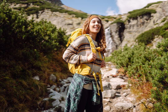 Female hiker with yellow backpack walks along mountain trail alongside stream. Woman enjoys  mountain scenery and feels a sense of freedom. Concepts of adventure, hiking, and sports.