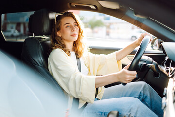 Plakat A young woman drives through a busy area during the day. She smiles, looking out the window. Sunlight floods the car's interior, creating a warm atmosphere. Urban and transportation concept.