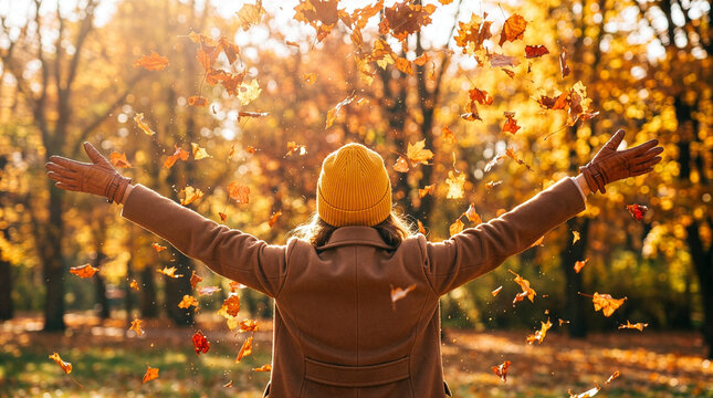 Joyful woman in brown coat and yellow hat throwing autumn leaves in the air with arms outstretched in a park during golden hour