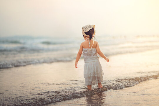 Adorable Asian toddler girl in a white dress exploring the beach shoreline at sunset golden hour. She stands peacefully watching ocean waves during a summer family travel holiday, capturing childhood