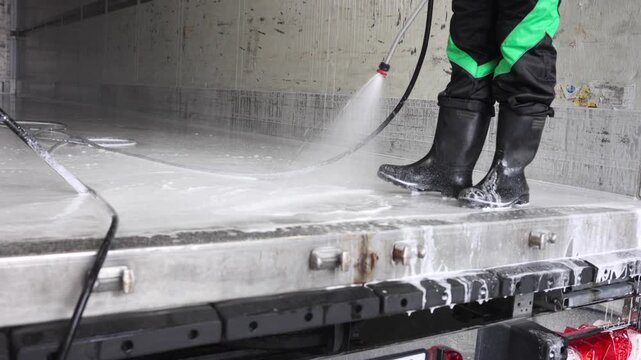 A uniformed cleaner washes the body of a large truck with a powerful jet of water with foam in rubber boots