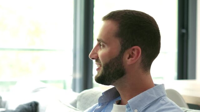Handsome bearded young man relaxing on a couch, looking out of a bright window, then turning to smile charmingly at the camera in a modern, naturally lit living room environment