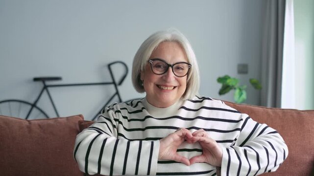 Happy senior woman with gray hair smiling and showing a heart gesture with her hands, expressing love and kindness while sitting on the sofa in her living room
