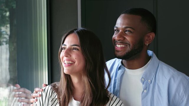 Young mixed-race couple standing by the window of their new home, looking outside with smiles and dreaming about the future together while sharing an intimate and affectionate moment