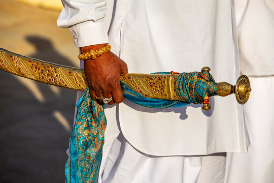 Man in traditional white attire holding a ceremonial golden sword. Close up of an indian man hand with gold jewelry gripping an ornate golden tulwar sword scabbard wrapped in blue silk scarf, India