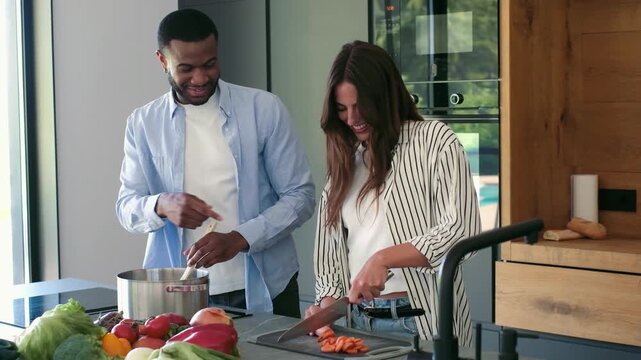Joyful multiethnic couple having fun while preparing a healthy meal with fresh vegetables in their modern kitchen, the woman chopping carrots while the man stirs a pot on the stove