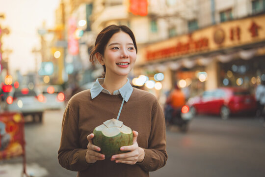 Happy young Asian woman traveler holding fresh coconut drink at chinatown street market Bangkok looking away with cheerful smile tourist exploring local food and culture urban lifestyle, Thailand