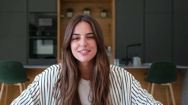 Happy young female vlogger looking at the camera and talking, actively gesticulating during a video conference while sitting in her modern kitchen. Remote work and online communication concept