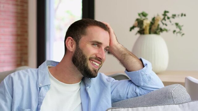 Young handsome man with a beard and blue eyes smiling while relaxing on the couch at home, daydreaming and looking away with a thoughtful and happy expression on his face