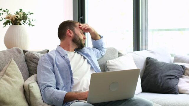 Frustrated young man feeling stressed and anxious while working on a laptop computer from his living room sofa, experiencing headache, burnout, and professional problems at his remote job