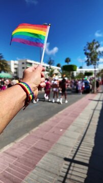 Close-up of a person's hand holding and waving a small rainbow flag during a sunny pride parade. The subject is wearing a rainbow bracelet, and the crowd is visible in the background