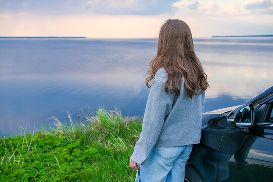 Woman standing near car and enjoying calm lake view during road trip before summer rainstorm
