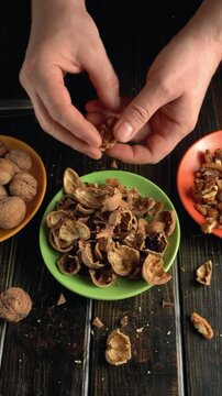 Hands cracking open walnut shells over a green bowl filled with empty shells, surrounded by whole walnuts and a bowl of walnut pieces