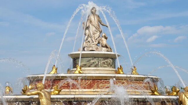 A medium shot captures the ornate tiered marble structure of the Latona
Fountain operating in the historical gardens of the Palace of Versailles. High
water arches spray from the top statue 
