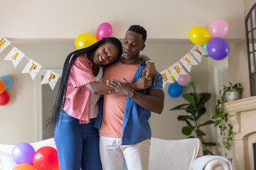 Fototapeta premium African American couple standing and hugging in home living room with balloons, bunting, couch