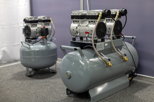 Industrial air compressors in equipment room. Two gray air compressors with metal tanks, hoses and motors displayed indoors for industrial, dental or workshop equipment use.