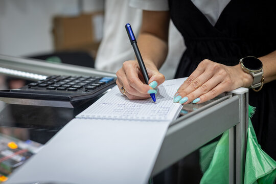Cashier Writing Receipt at Checkout Counter. Close-up of a shop worker writing notes on paper beside a cash register at a retail checkout counter.