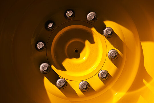 Yellow machinery wheel rim showing lug nuts, painted metal surfaces and strong shadow patterns. Industrial texture, mechanical design, durability and heavy equipment detail.