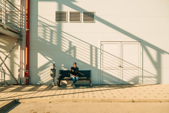 Woman sitting on bench in sunlit urban setting with dramatic staircase shadows and closed door