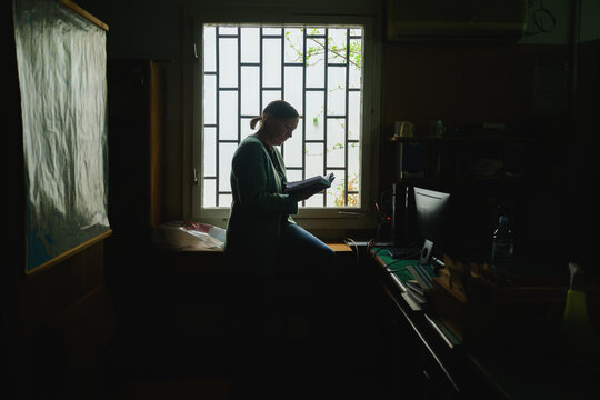 Female librarian standing by window in old library office reading a book, soft daylight and quiet academic atmosphere