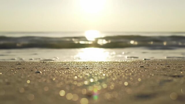 Extreme low angle close-up video of gentle ocean waves with white foam edges washing a pristine golden sandy beach at golden hour, with sun flares and reflections. Peaceful twilight aura