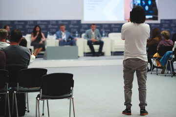 Fototapeta premium Person stands facing the conference panelists and records video on a smartphone. Moderator faces group of seated speakers. Audience watches attentively.
