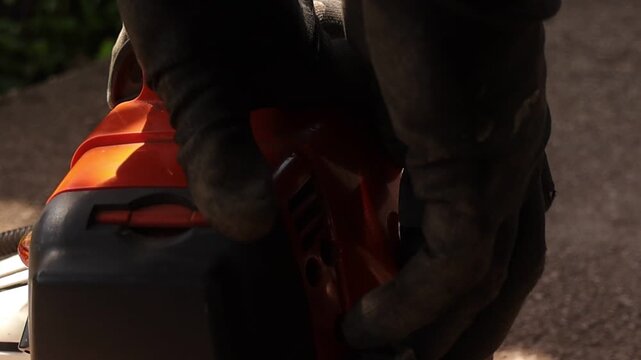 Detailed close up of hands pulling the starter cord on a motorized grass trimmer during lawn care preparation, with focus on engine, recoil starter and protective gloves in sunlight.